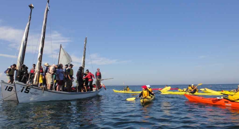 a group of people stand in a sailboat. There are others paddling colorful kayaks nearby. 